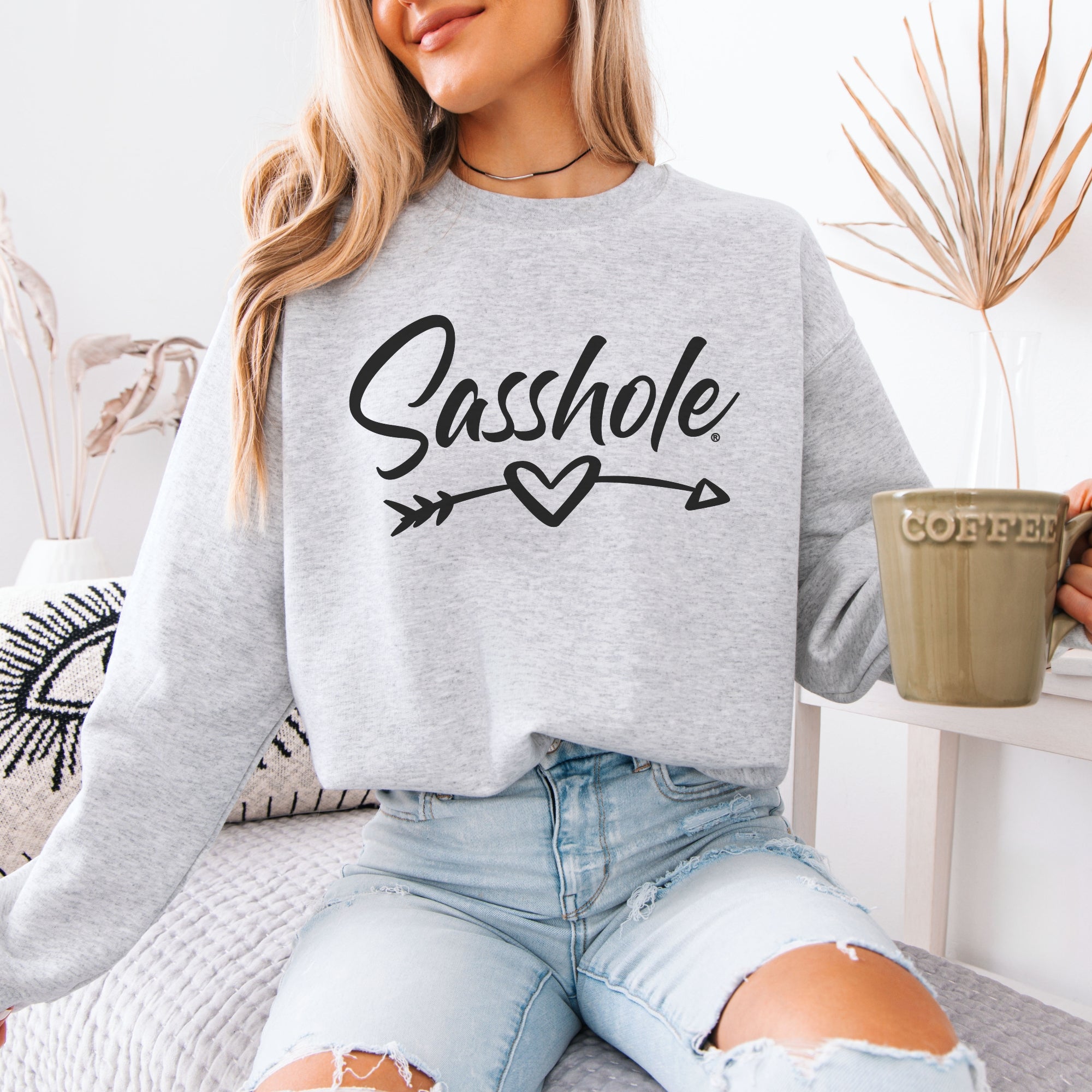 A woman sits comfortably, holding a "Coffee" mug and wearing a "sasshole" graphic sweatshirt and ripped denim jeans.

