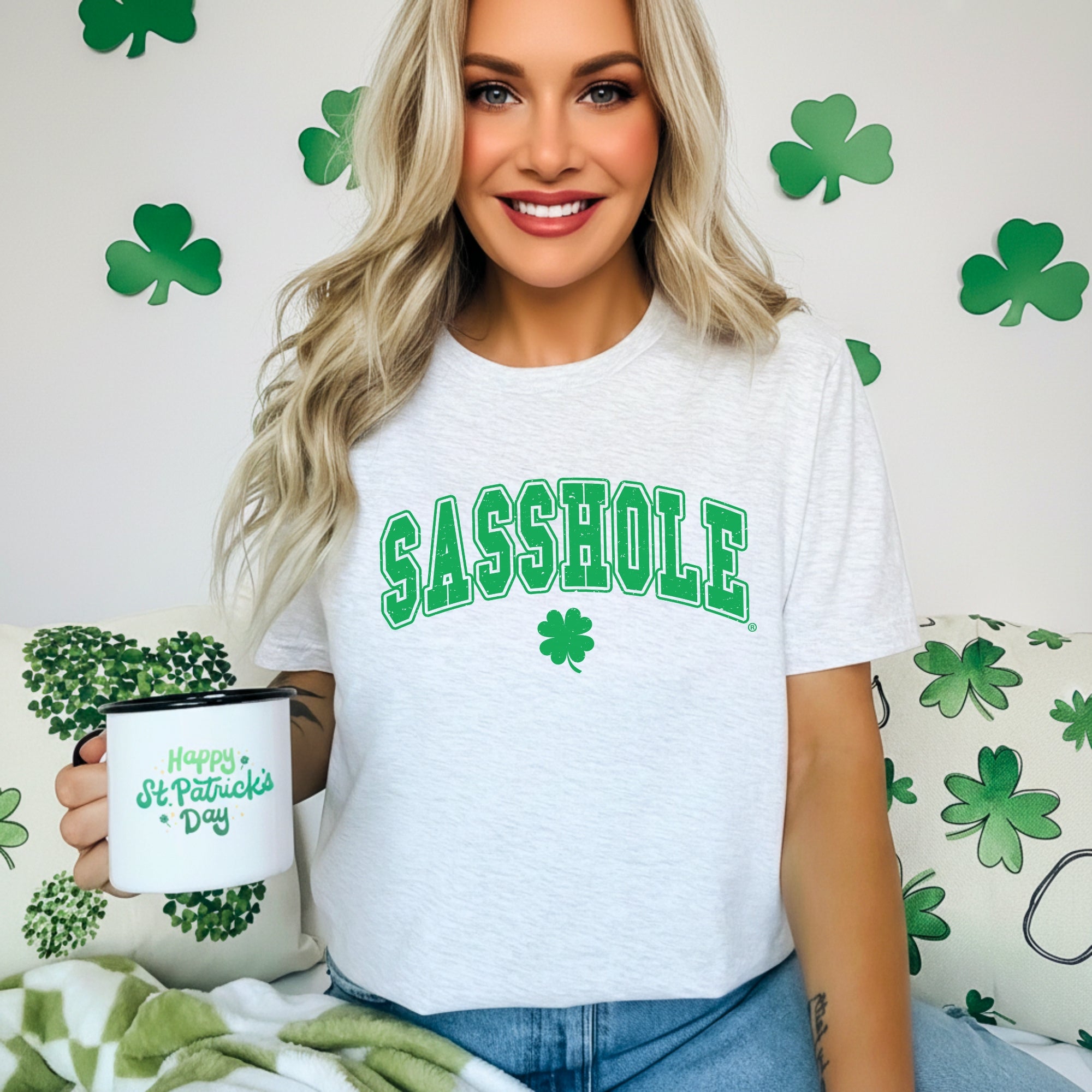 Image of Woman wearing a white t-shirt with 'SASSHOLE' in green text, holding a mug with St. Patrick's Day design, against a shamrock patterned background.
