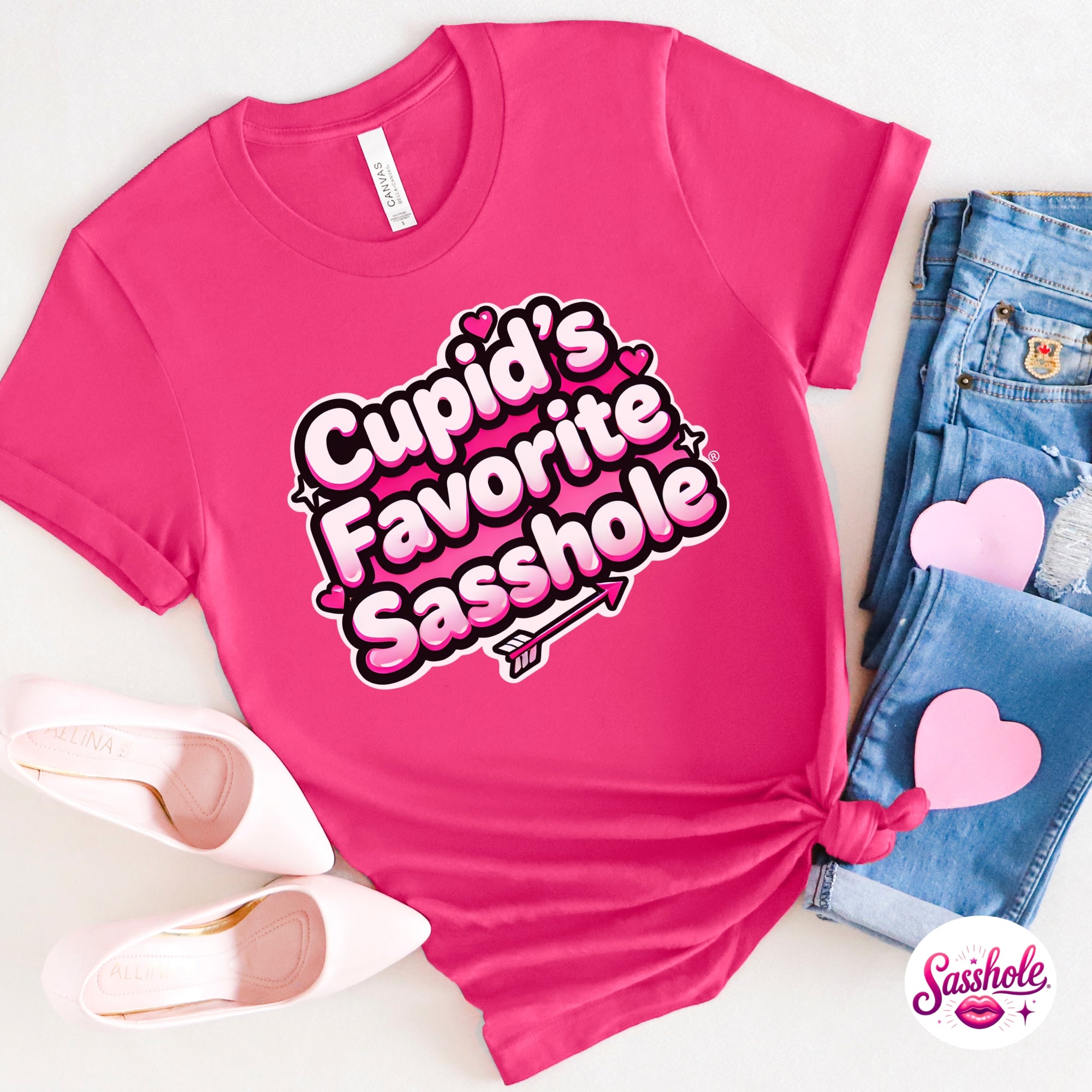 Pink t-shirt with 'Cupid's Favorite Sasshole' text, surrounded by denim jeans and pink shoes on a white background.