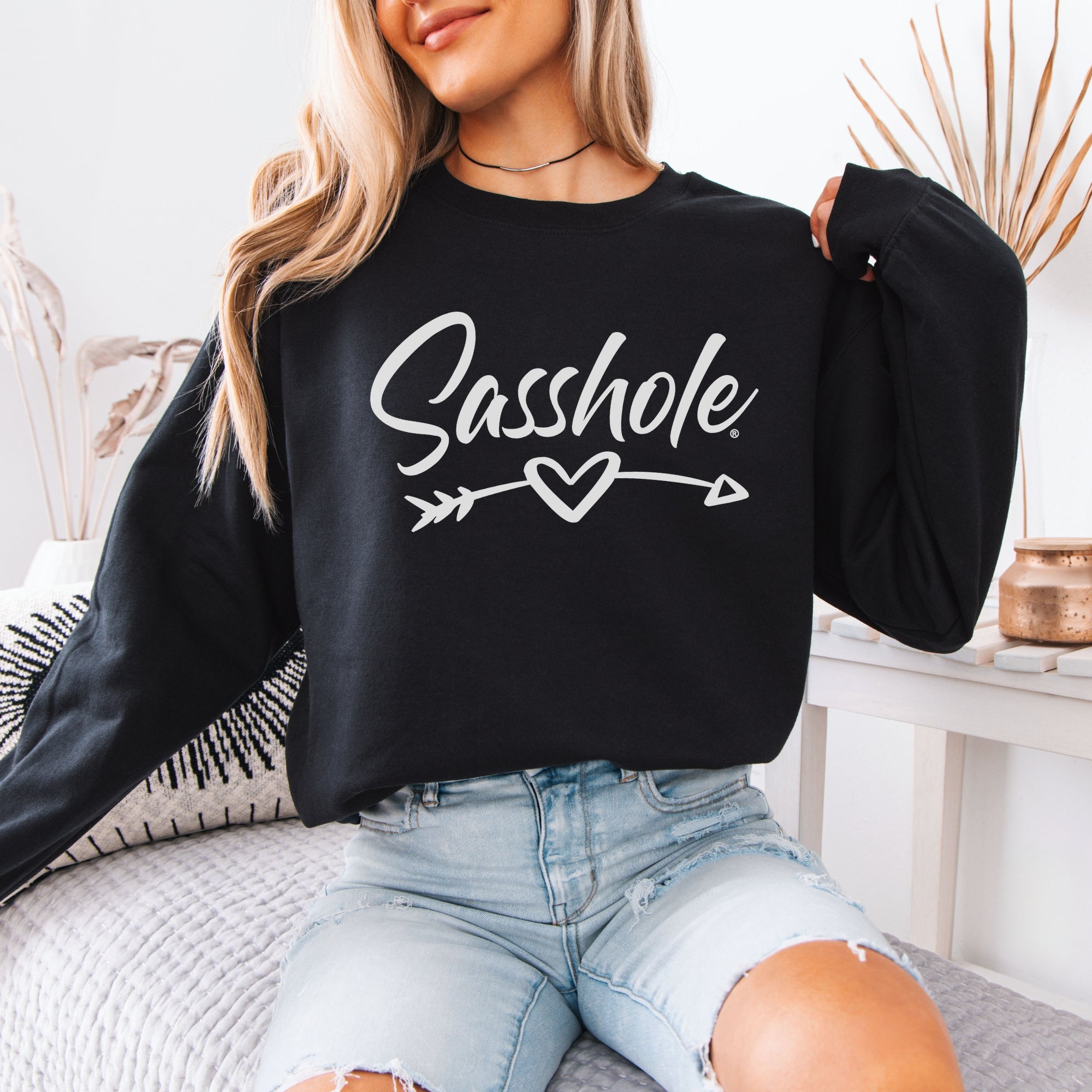 A woman models a black "Sasshole" sweatshirt with a heart arrow design while sitting on a bed in denim shorts.
