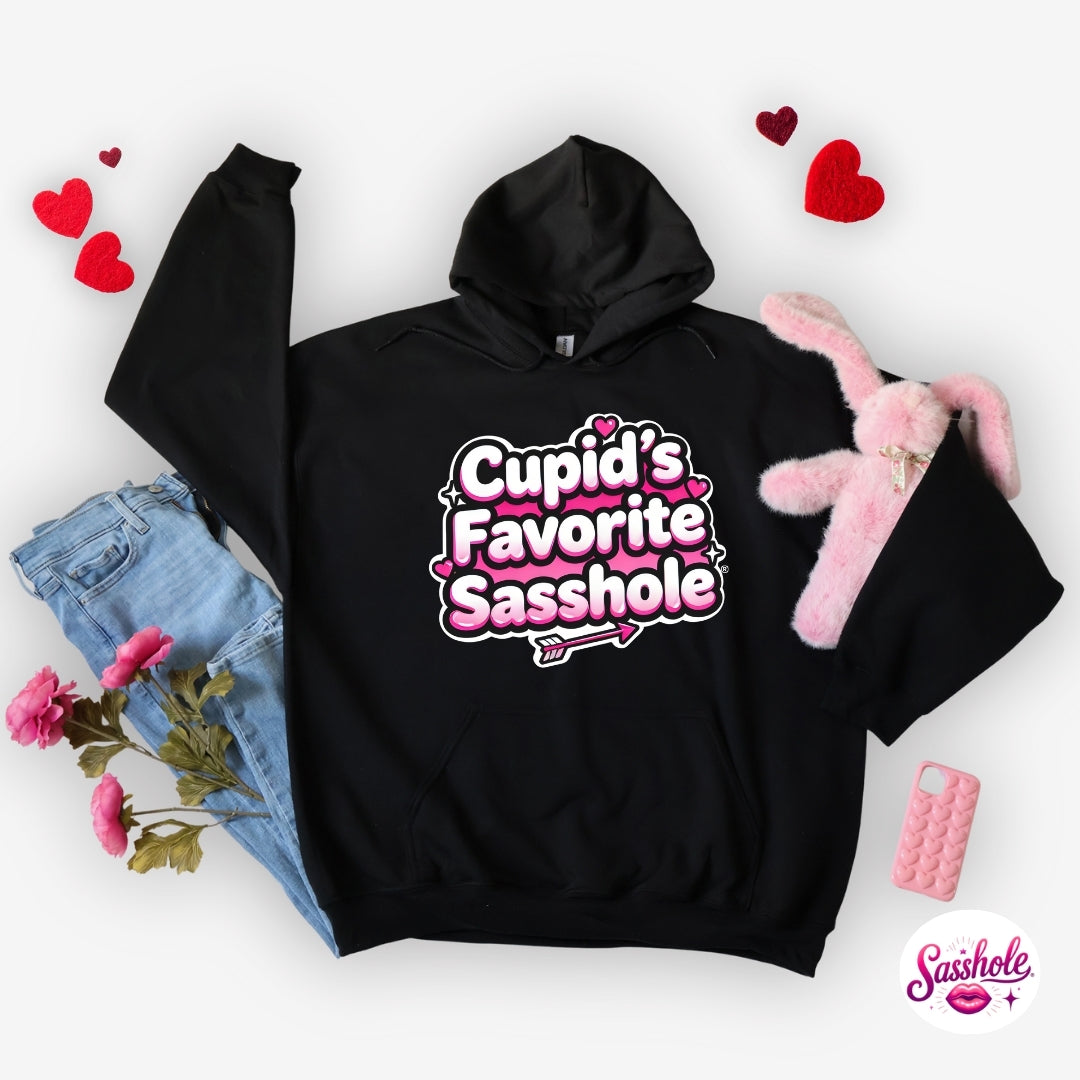 Image of Black hoodie with 'Cupid's Favorite Sasshole' text, surrounded by jeans, flowers, and a pink plush toy on a white background.