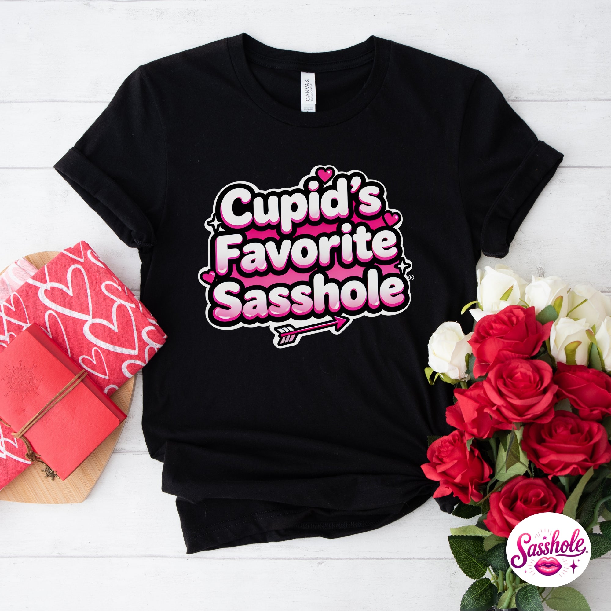 Black t-shirt with 'Cupid's Favorite Sasshole' text, surrounded by flowers and a gift box on a white background.