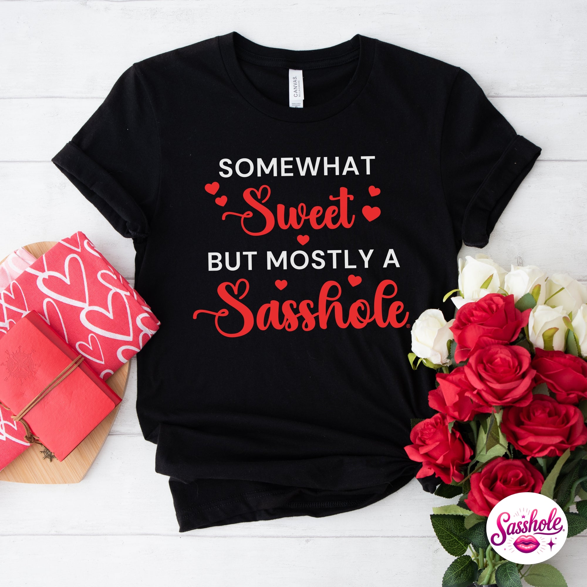 Black t-shirt with humorous text design on a white wooden surface with flowers and gift boxes.