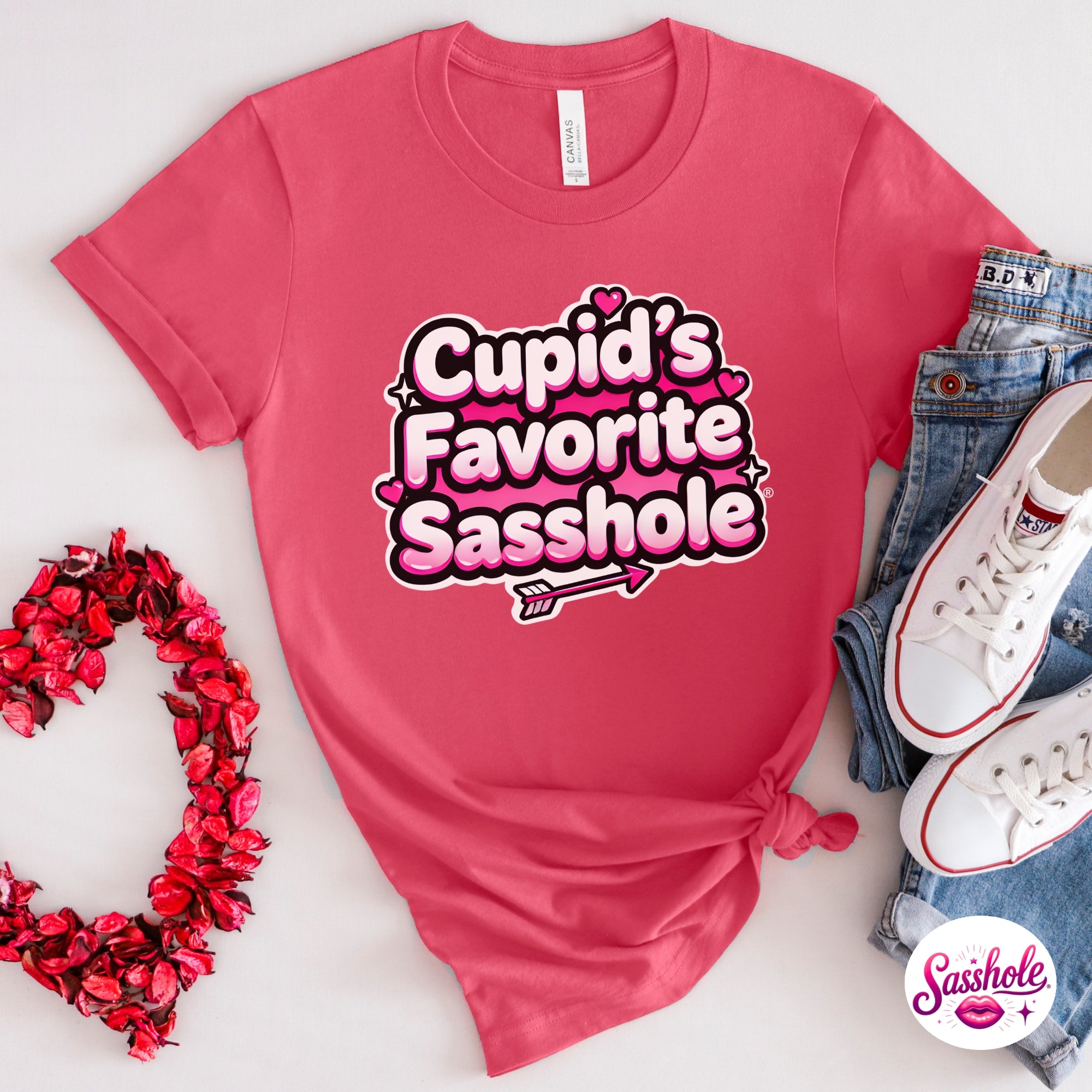 Pink t-shirt with 'Cupid's Favorite Sasshole' text, surrounded by denim jeans, white sneakers, and a heart-shaped decorative item.
