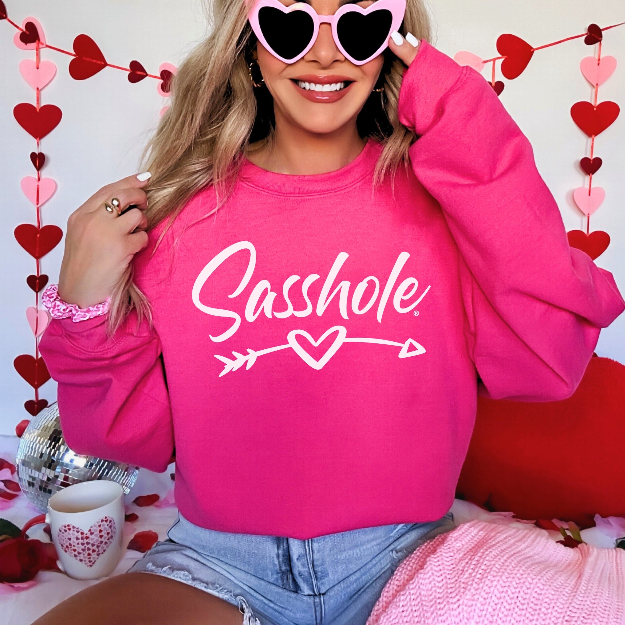 Person wearing a pink sweatshirt with 'Sasshole' text, sitting against a heart-themed background.
