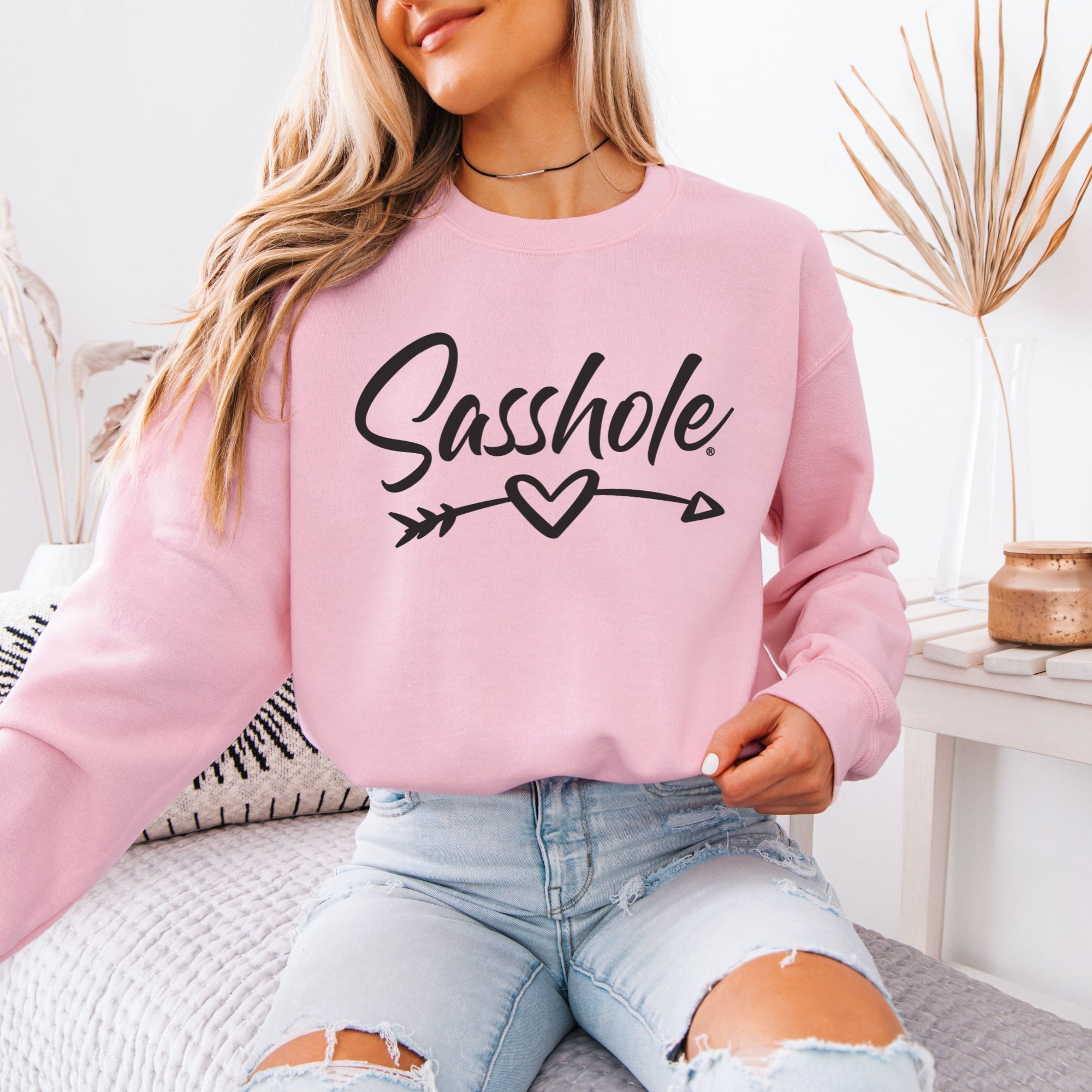 Woman models a light pink "Sasshole" sweatshirt with ripped jeans in a bohemian-style setting.
