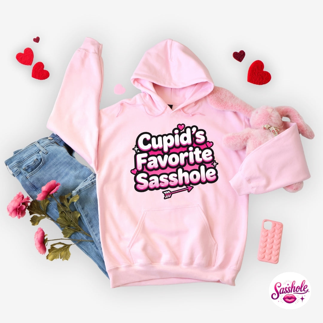 Image of Pink hoodie with 'Cupid's Favorite Sasshole' text, surrounded by jeans, flowers, and a phone case on a light background.