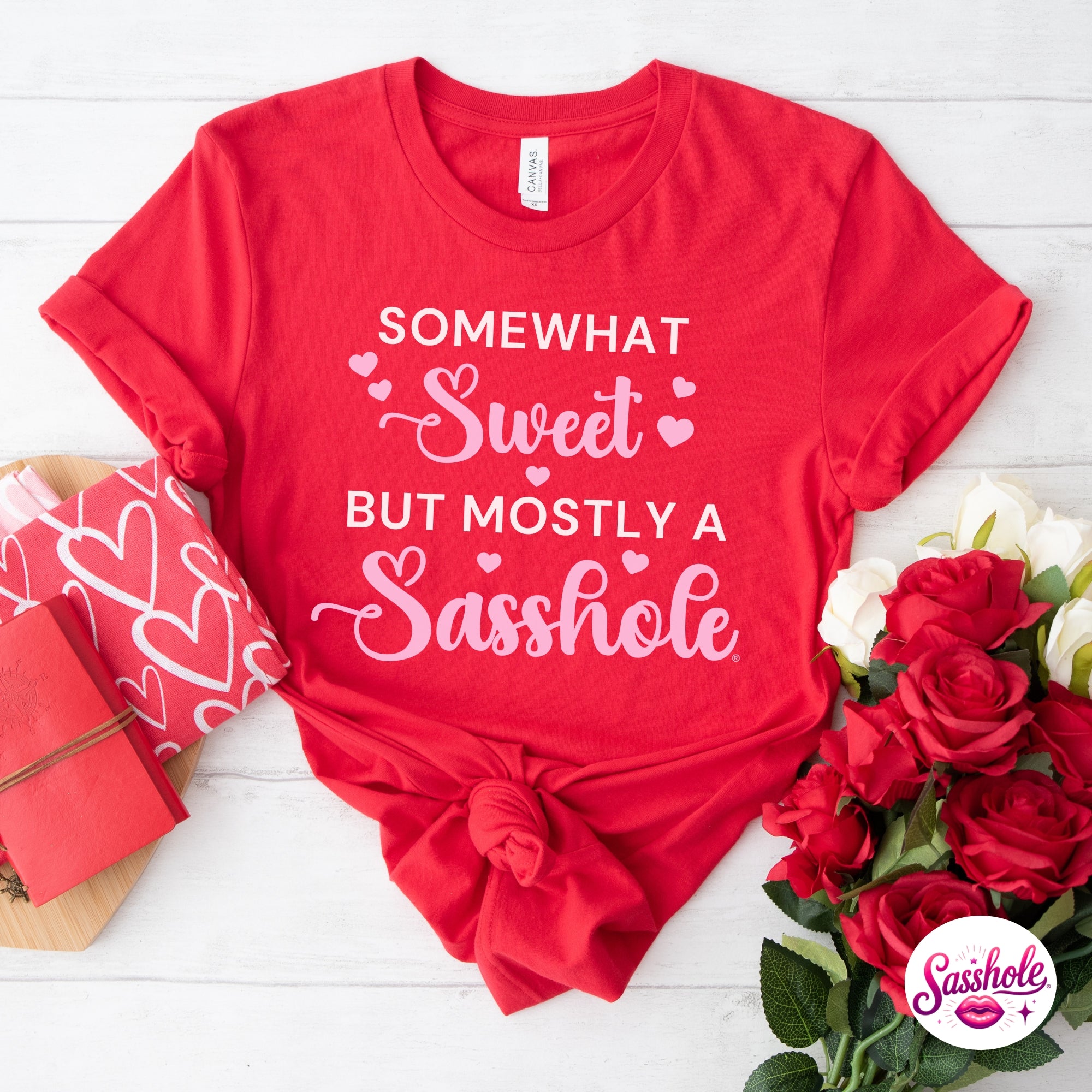 Red t-shirt with humorous text on a white wooden surface with flowers and gift box.