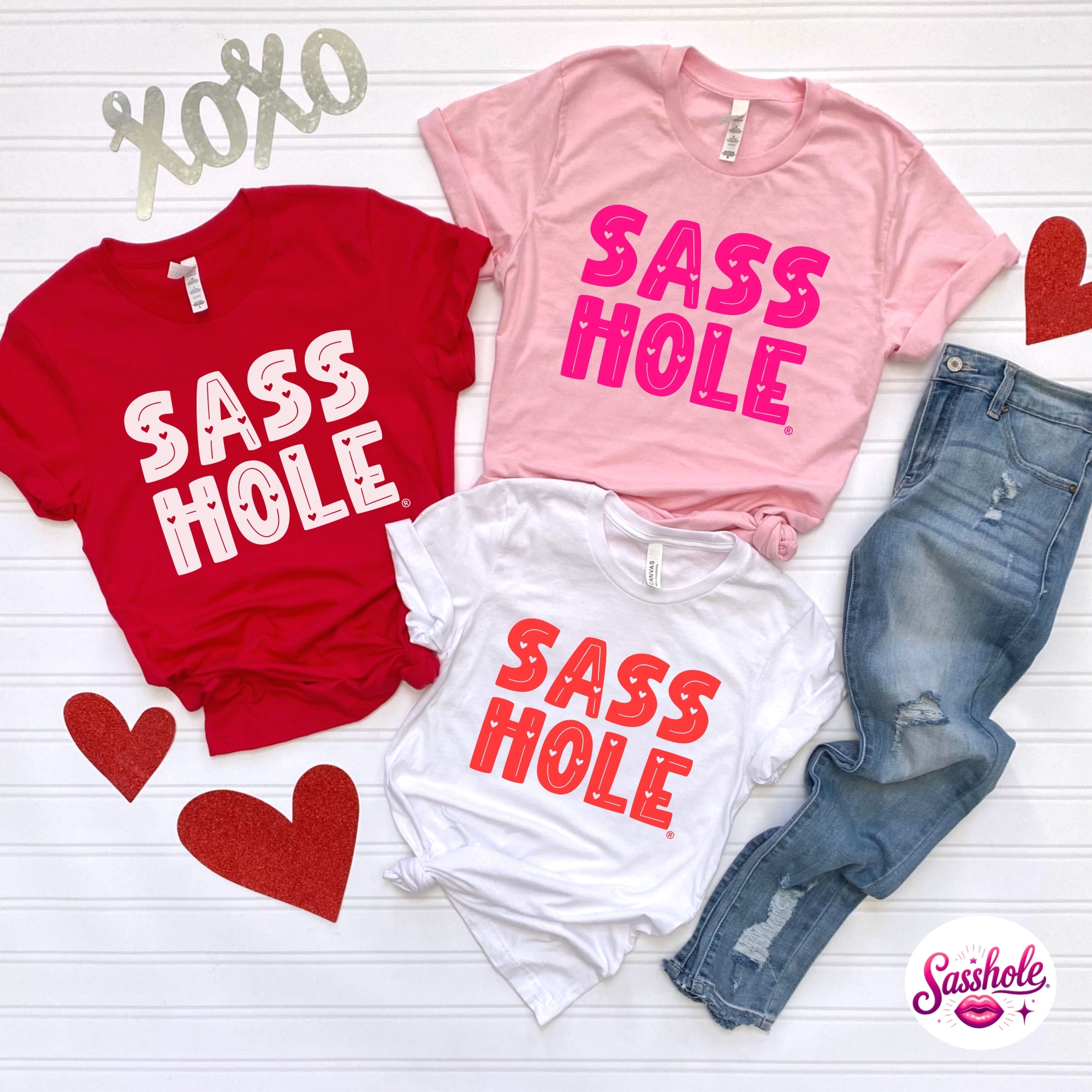 Three shirts with 'SASS HOLE' text on a white surface with jeans and hearts.