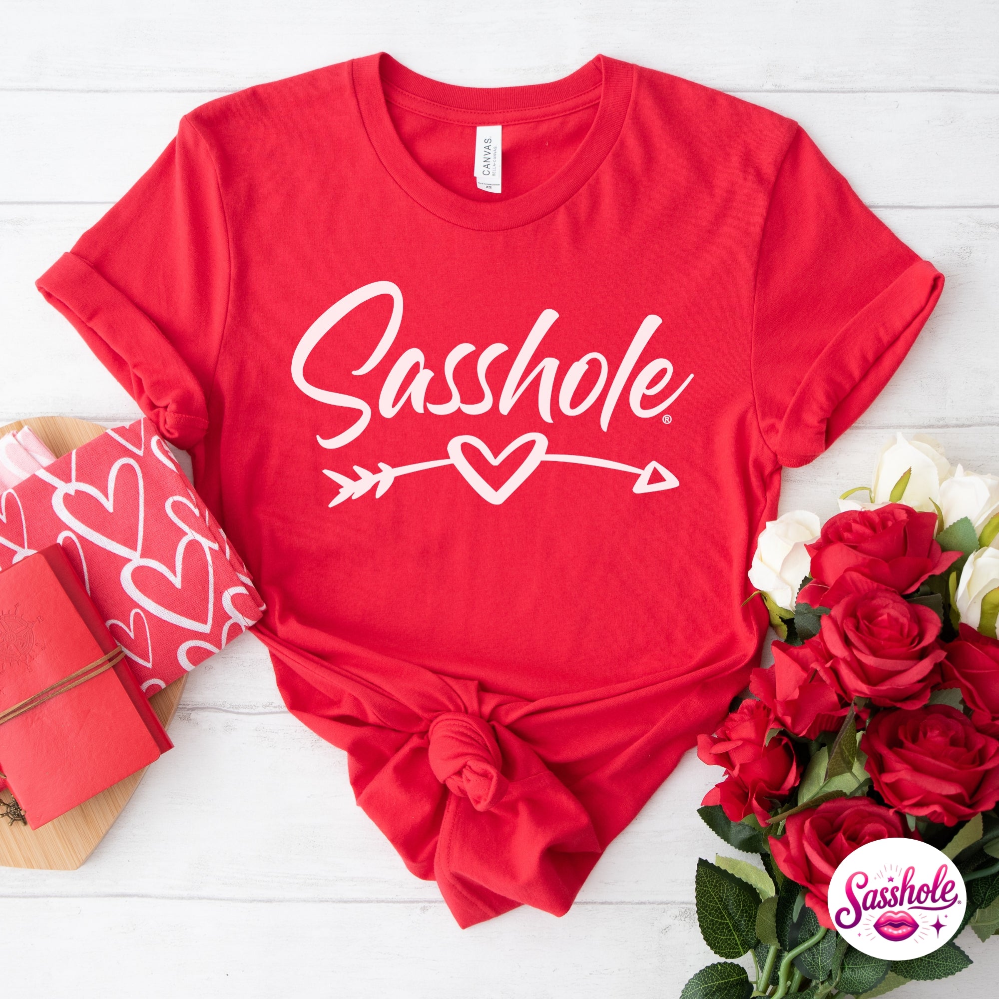 Image of Red t-shirt with 'Sasshole' text on a white wooden surface with flowers and gift box.