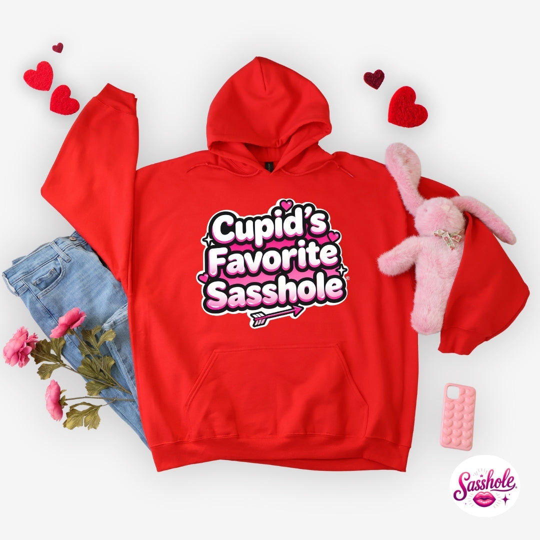 Image of Red hoodie with 'Cupid's Favorite Sasshole' text, surrounded by denim jeans, a pink plush toy, and heart-shaped decorations on a white background.