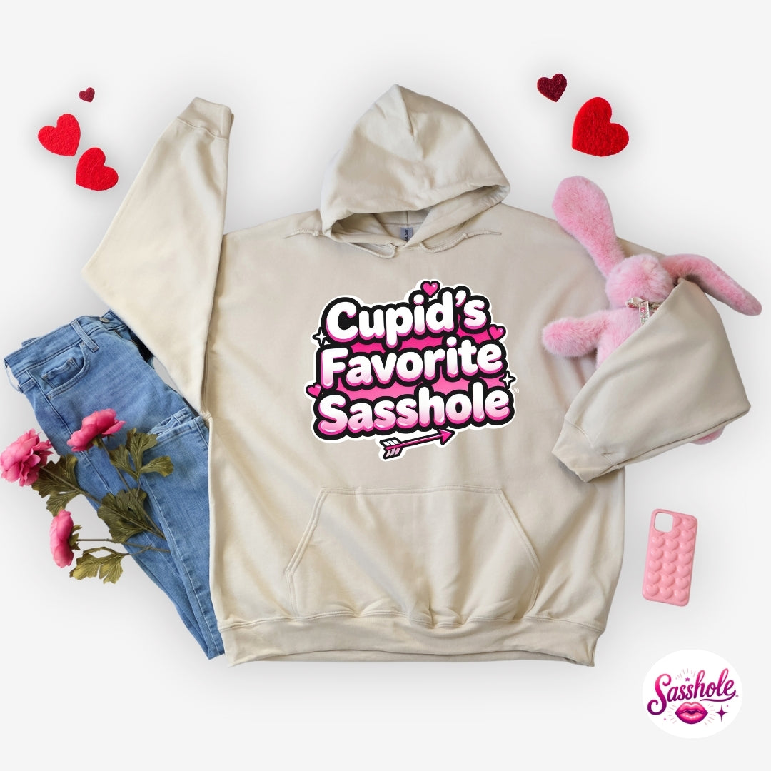 Beige hoodie with 'Cupid's Favorite Sasshole' text, surrounded by denim jeans, pink plush bunny, and decorative hearts on a white background.