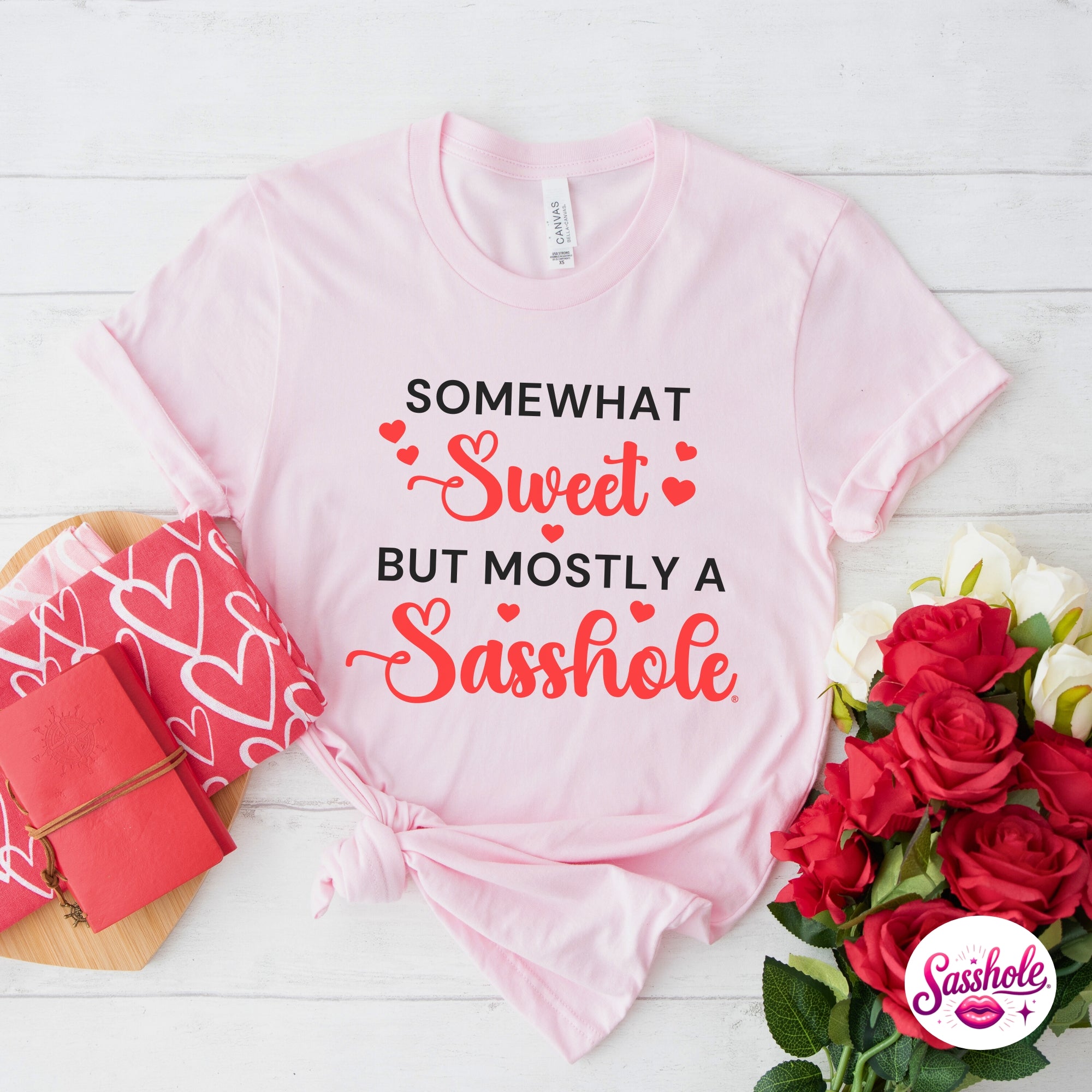 Image of Pink t-shirt with humorous text design on a white wooden surface with flowers and gift box.