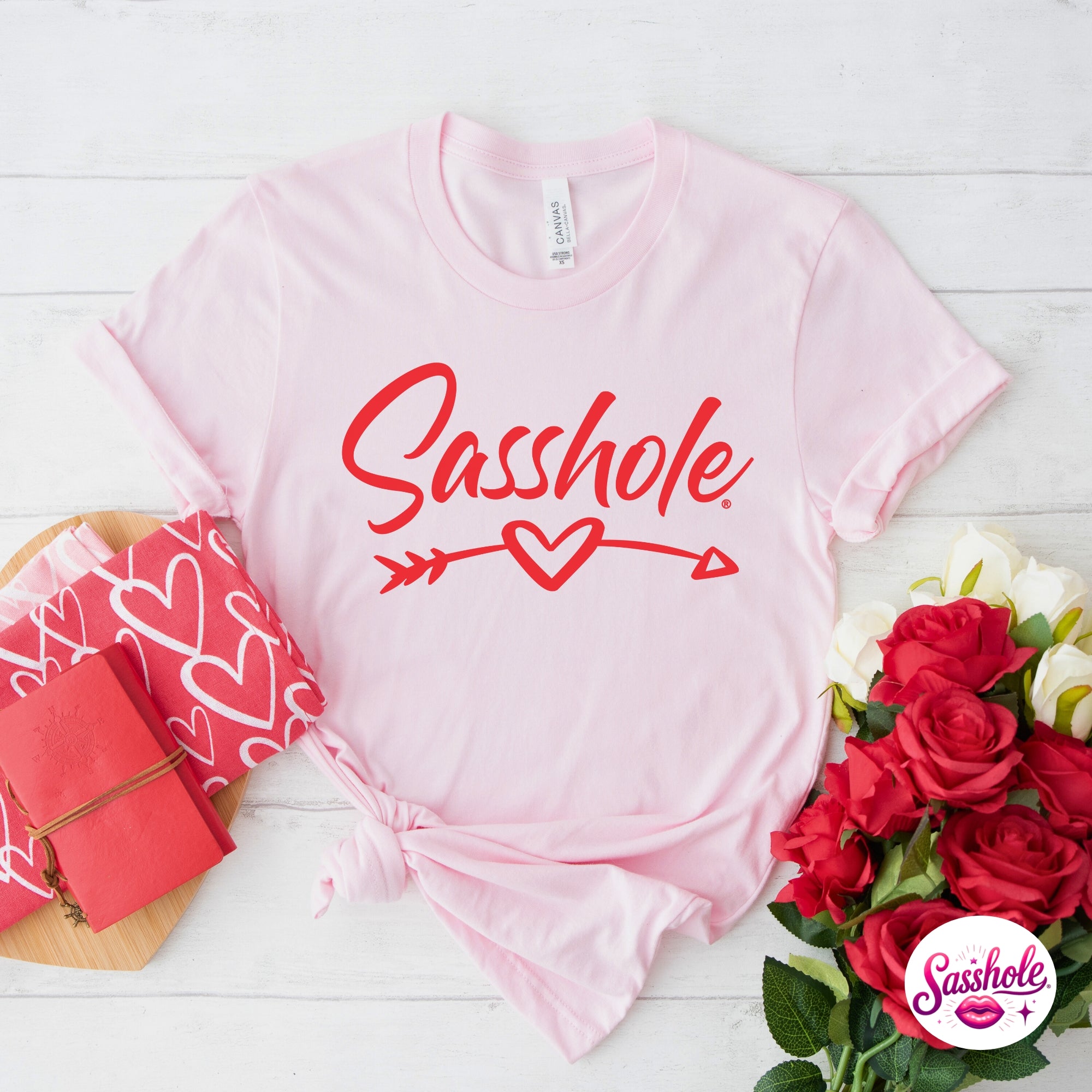 Pink t-shirt with 'Sasshole' text, red roses, and gift box on a white wooden surface.