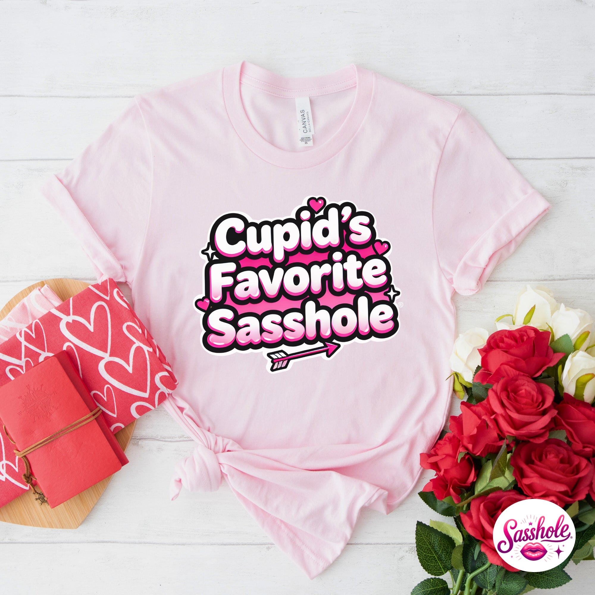 Pink t-shirt with 'Cupid's Favorite Sasshole' text, surrounded by roses and a gift box on a white wooden surface.