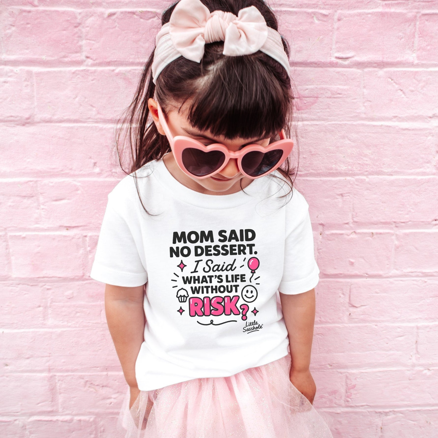 Child wearing a white t-shirt with a humorous quote, pink tutu, and heart-shaped sunglasses against a pink brick wall.