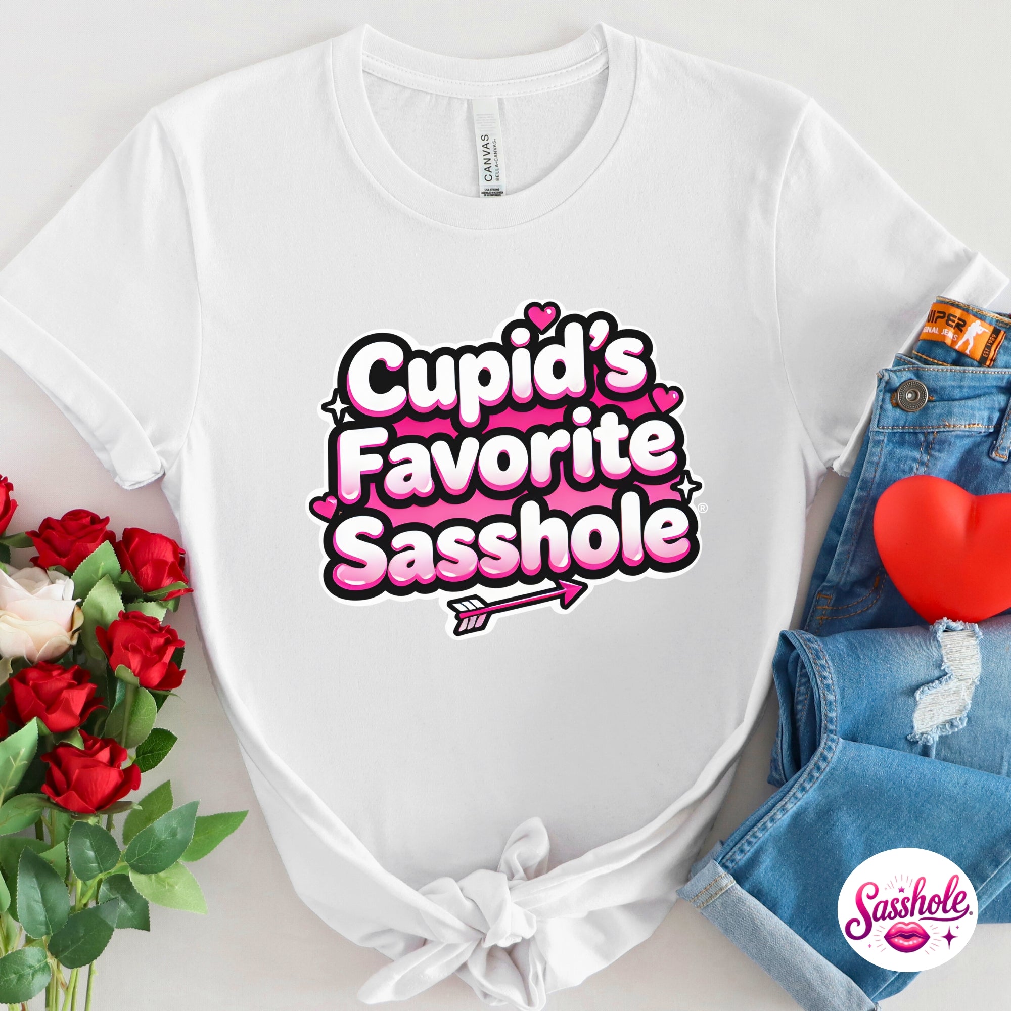 White t-shirt with 'Cupid's Favorite Sasshole' text, surrounded by flowers and denim jeans on a light gray background.