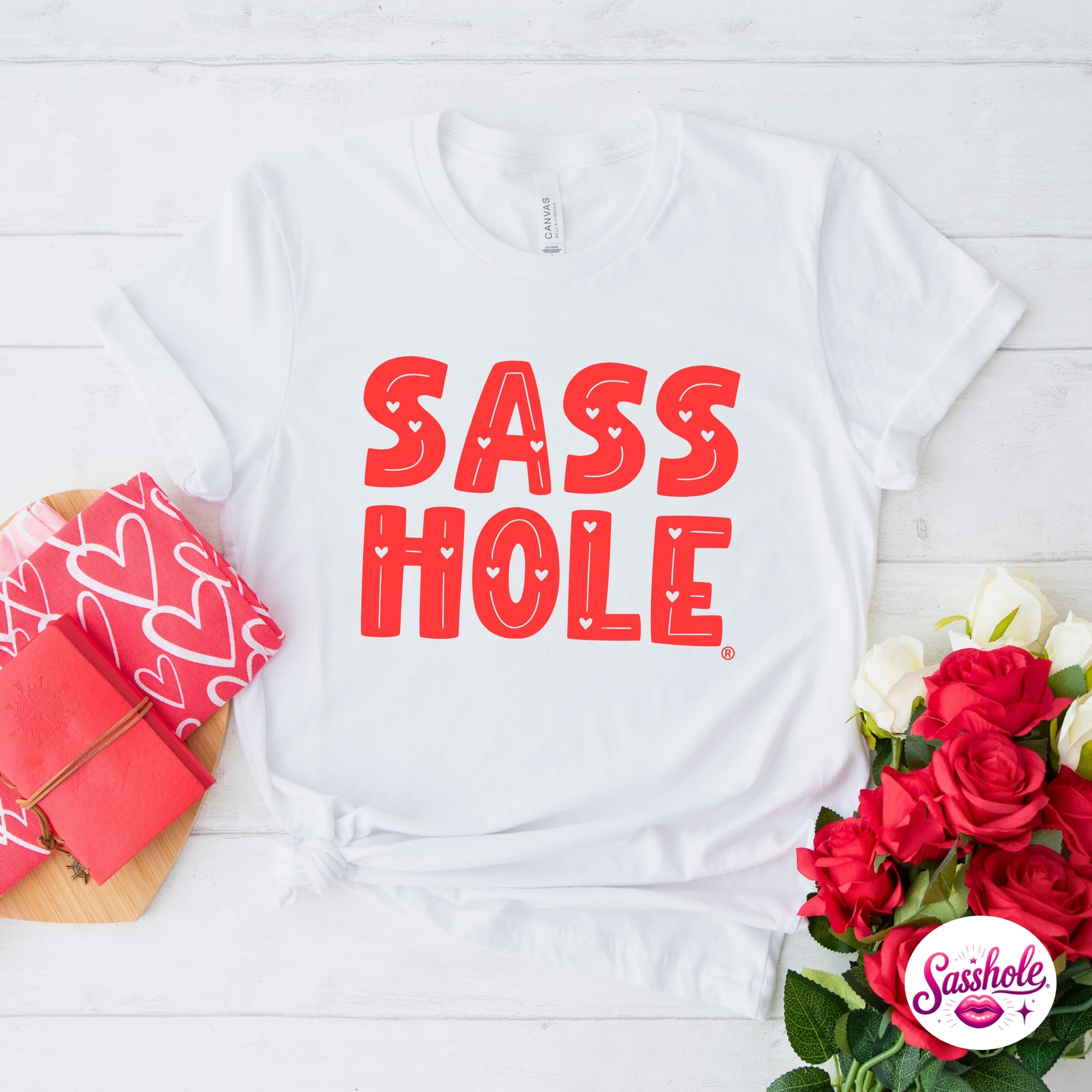 White t-shirt with 'SASS HOLE' in red text, surrounded by red roses and a pink gift box on a light wooden surface.