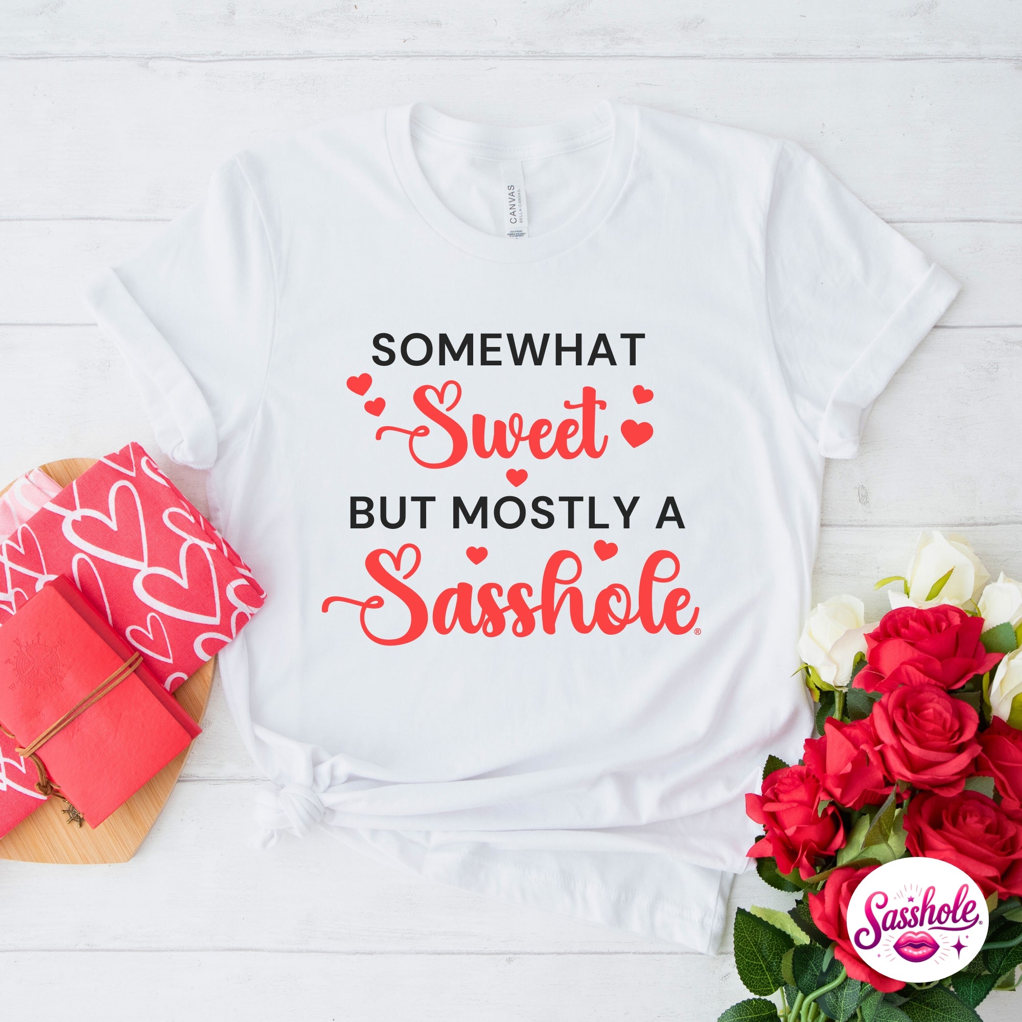 Image of White t-shirt with humorous text design on a white wooden surface with flowers and a pink box.