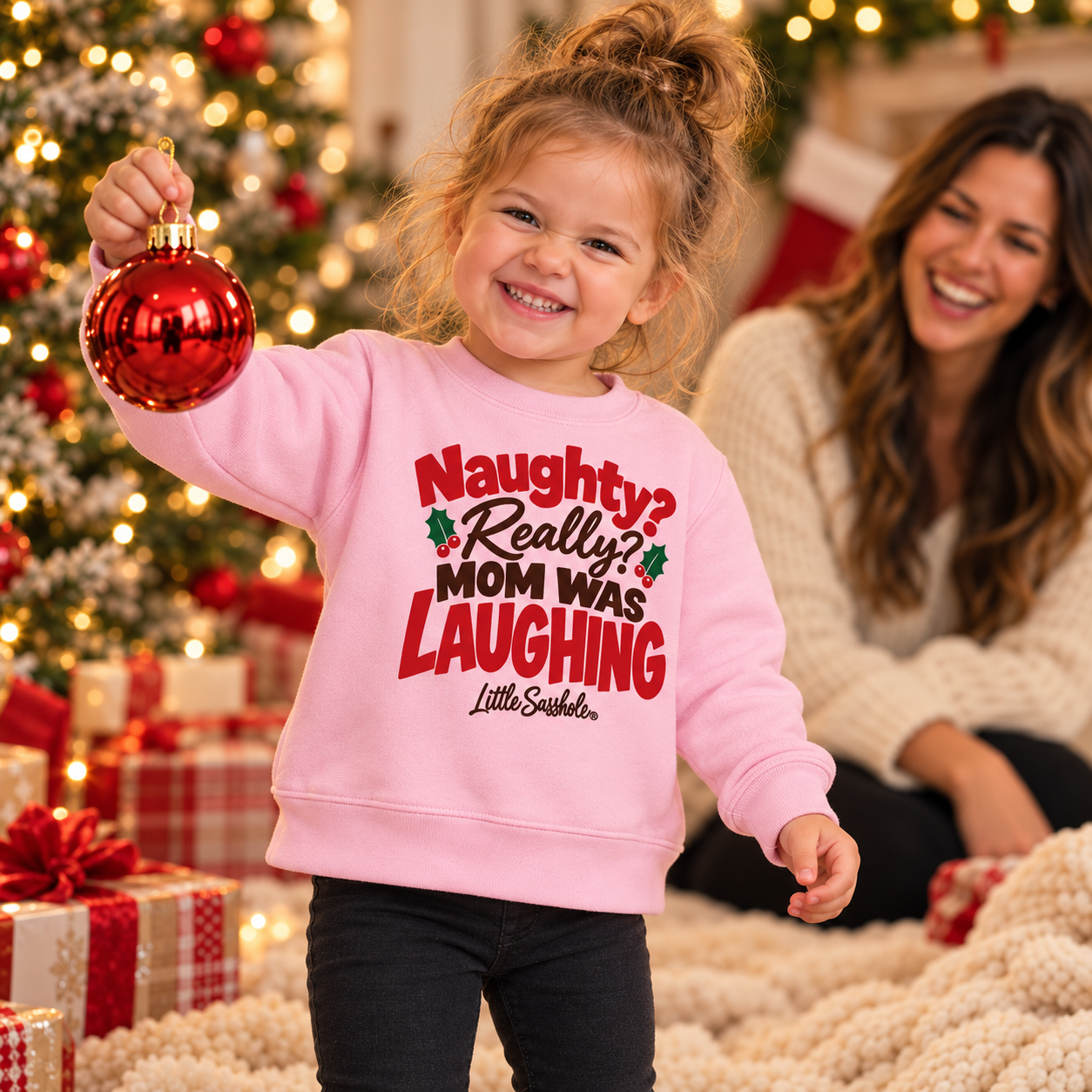 Image of Toddler wearing the Pink Mom Was Laughing Christmas sweatshirt and holding a red ornament beside her mother.