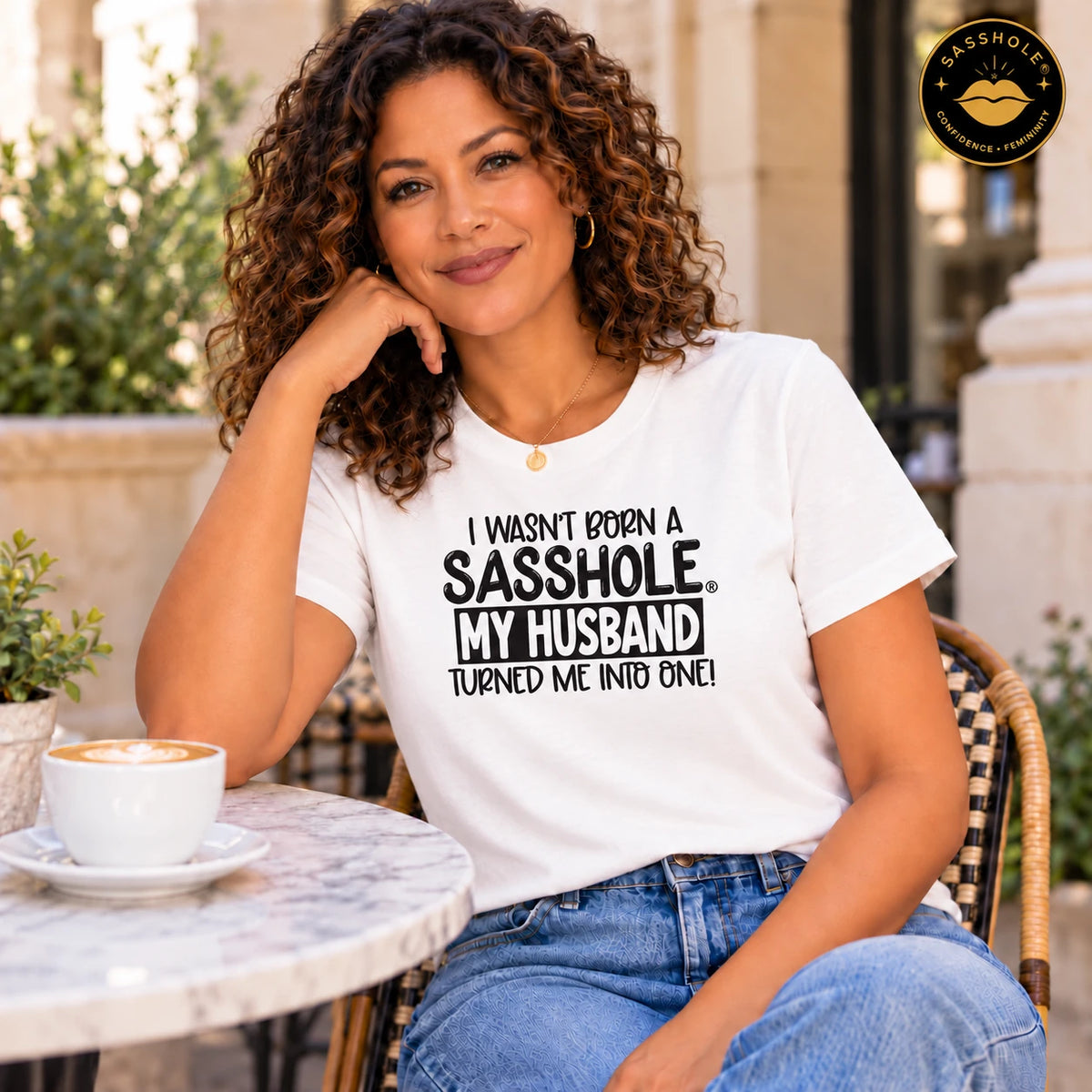 I Wasn’t Born a Sasshole, My Husband Turned Me Into One! tee in White worn at an outdoor cafe.